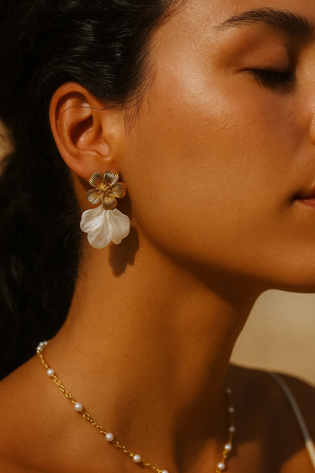 Nahaufnahme des Gesichtsprofils einer jungen Frau mit gebräunter Haut, die gold-weiße Blumenohrringe und eine feine goldene Halskette mit weißen Perlen trägt. Das warme, natürliche Licht betont ihre Hauttextur und die Details des Schmucks vor einem dezenten Hintergrund.