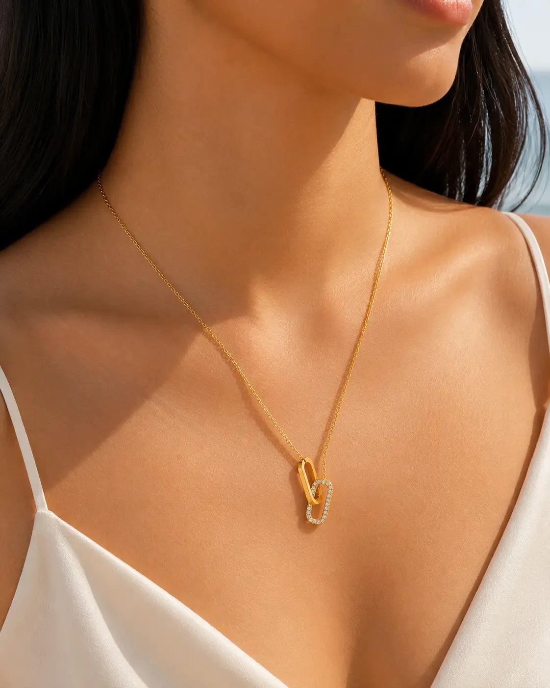 Goldene Halskette mit zwei ineinandergreifenden Anhängern, davon einer mit funkelnden Steinen besetzt, am Dekolleté eines dunkelhaarigen Models im warmen Sommerlicht.
