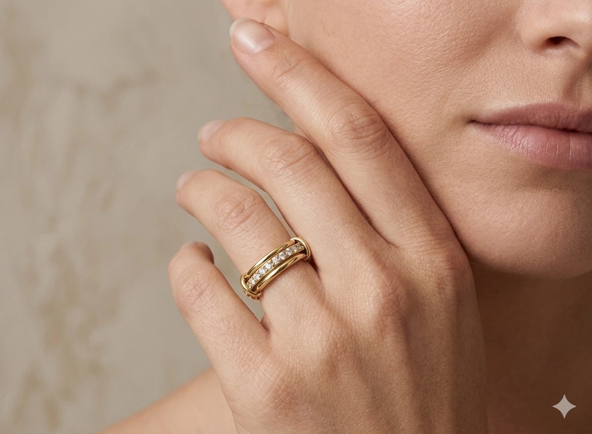 Varalida Ring - Elegante Lifestyle-Nahaufnahme eines luxuriösen Goldrings mit Diamantbesatz, getragen am Ringfinger einer Frau. Die Hand ruht sanft an der Wange vor einem weichen, beigen Hintergrund. Der Ring zeichnet sich durch ein dreiteiliges Design aus: zwei polierte Goldbänder umschließen eine mittlere Schiene, die mit funkelnden Zirkonia- oder Diamantsteinen besetzt ist. Das Licht setzt feine Akzente auf das Metall und die Steine, was die hochwertige Verarbeitung des Schmuckstücks unterstreicht.
