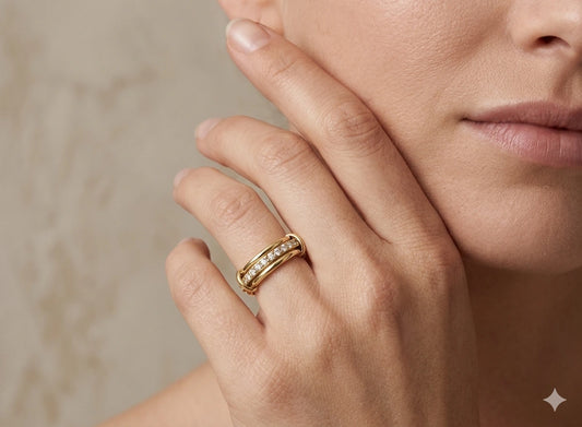 Varalida Ring - Elegante Lifestyle-Nahaufnahme eines luxuriösen Goldrings mit Diamantbesatz, getragen am Ringfinger einer Frau. Die Hand ruht sanft an der Wange vor einem weichen, beigen Hintergrund. Der Ring zeichnet sich durch ein dreiteiliges Design aus: zwei polierte Goldbänder umschließen eine mittlere Schiene, die mit funkelnden Zirkonia- oder Diamantsteinen besetzt ist. Das Licht setzt feine Akzente auf das Metall und die Steine, was die hochwertige Verarbeitung des Schmuckstücks unterstreicht.